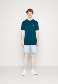 Camiseta de manga corta en color teal con un cuello redondo y un pequeño logo amarillo en el pecho, combinada con pantalones cortos de mezclilla azul claro y zapatillas blancas.