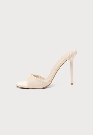 Beige muil met hoge hak en een smalle stilettokurk en een open teen, tegen een effen lichtgrijze achtergrond.