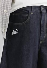 Jean en denim bleu foncé avec un texte brodé blanc près de la poche avant, porté avec une chemise blanche et une chemise à carreaux gris clair boutonnée.