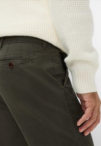 Pantaloni di cotone verde oliva scuro con una tasca posteriore dotata di un bottone marrone, abbinati a un maglione beige strutturato.