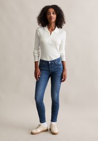 Biała prążkowana koszulka polo z długim rękawem, niebieskie dopasowane dżinsy i beżowe sneakersy z teksturowaną podeszwą, na neutralnym tle.