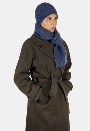 Frau trägt einen dunkelbraunen Mantel mit Gürtel, eine blaue Strickmütze und einen passenden blauen Schal, steht mit den Händen in den Manteltaschen und schaut nach rechts.