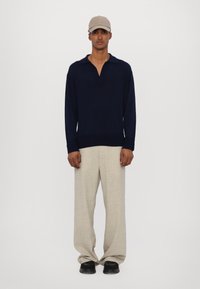 Tmavě modré tričko s dlouhým rukávem a límečkem, světlé béžové široké kalhoty a černé boty; osoba má na sobě béžovou čepici. Jednoduchý, čistý design.