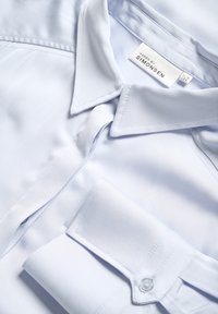 Cămașă bleu deschis, cu nasturi, dintr-un material neted, având guler ascuțit, manșon unic și o etichetă care spune „Karen by Simonsen.”