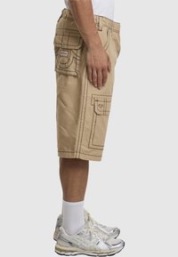 Beige Cargo-Shorts mit mehreren Taschen, kontrastierenden dunklen Nähten, entspannter Passform und knielangem Schnitt, kombiniert mit silbernen Sportschuhen.