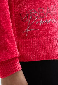 Roter strukturierter Pullover mit langen Ärmeln, verziert mit schwarzen Nieten und silberner Schrift auf der Brust, dargestellt an einem Arm und Torso einer Person.