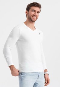 Maglietta a maniche lunghe bianca con scollatura a V, realizzata in tessuto morbido, con un piccolo logo sul petto e un design aderente. Abbinata a jeans azzurri chiari.