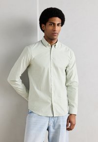 Νεαρός άνδρας με κοντά αφρο μαλλιά, που φοράει ένα πουκάμισο με κουμπιά σε ανοιχτό πράσινο και λευκές ρίγες και ανοιχτό μπλε τζιν, με φόντο έναν απλό τοίχο.