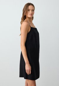 Robe en lin noir à bretelles fines, décolleté carré et coupe fluide. Présente des coutures minimales et aucun motif supplémentaire.