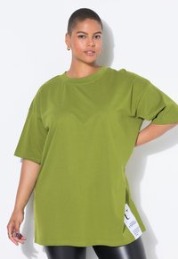 Camiseta de algodón de color verde oliva, de corte oversize, con hombros caídos, mangas cortas y detalle de etiqueta lateral. Textura suave y diseño casual.