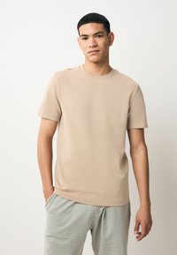 T-shirt beige à manches courtes avec un col rond, une texture lisse et une coupe décontractée, associé à un pantalon rayé vert et blanc.