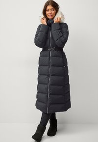 Long manteau rembourré noir avec une taille ceinturée, doté d'une fermeture éclair à l'avant et d'une capuche bordée de fausse fourrure. Design texturé et matelassé.