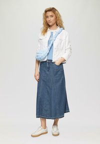 Lichtblauwe crossbody tas met een gedraaid ontwerp, witte denimjas, gestreept blauw-wit shirt en een lange denimrok. Witte sneakers.