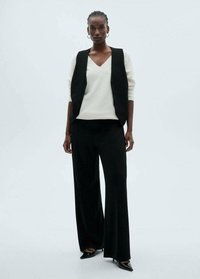 Vesta neagră peste un pulover alb cu decolteu în V, asortată cu pantaloni negri cu picioare largi și pantofi negri ascuțiți. Textură netedă, design minimalist.