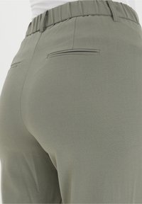 Närbild av person som bär ljus olivgröna byxor med elastiskt midjeband och två bakfickor med welt.