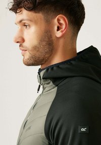 Herrenjacke in Oliv und Schwarz, mit gestepptem Material, Reißverschluss und Kapuze. Auffälliger Markenpatch am Ärmel.