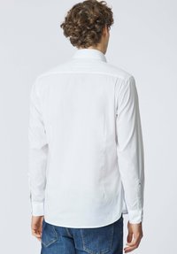 Uomo con capelli ricci che indossa una maglia bianca aderente a maniche lunghe e jeans blu, mostrato di spalle su uno sfondo semplice.