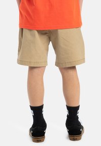 Pantalones cortos caqui con dobladillo, combinados con zapatillas negras y calcetines negros con un logo blanco. Camiseta naranja brillante visible en la parte superior.