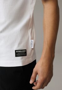 Weißes Baumwoll-T-Shirt mit einem schwarz gestickten Etikett mit der Aufschrift "STRELLSON" und einem kleineren weißen Tag. Der Stoff zeigt eine glatte Textur.
