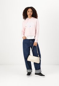 Cropped hoodie rosa con logo, jeans blu oversize e sneakers neri. Borsa crema dal design minimal. Texture del tessuto lisce.