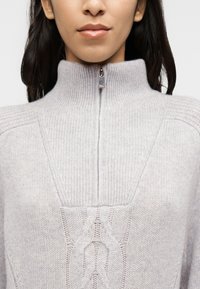Ανοιχτό γκρι πουλόβερ με ψηλό γιακά, κλείσιμο με μισή φερμουάρ και  διακοσμητική πλέξη κοτσίδα στο μπροστινό μέρος. Μαλακό, υφασμάτινο υλικό με υφή.