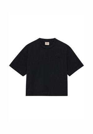 Kvinde med krøllet hår står med front mod kameraet iført en løs sort t-shirt, sorte cykelshorts, hvide sokker og beige vandresko.