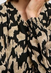 Tissu noir avec un motif abstrait beige, doté d'un décolleté en V profond et d'un détail de poignet froncé, mettant en valeur une texture lisse et un design fluide.