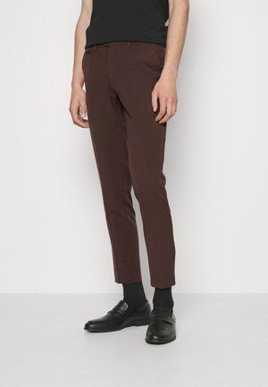 Pantaloni marroni su misura con vestibilità slim, texture liscia e pieghe pressate, abbinati a mocassini neri e calzini fino alla caviglia.