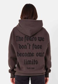 Sweat à capuche marron foncé avec un grand texte noir au dos. Doté de poignets et d'un ourlet côtelés, d'une texture douce et d'une coupe décontractée.