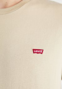 T-shirt in cotone beige con scollatura rotonda, caratterizzato da un logo Levi's ricamato in rosso sull'area sinistra del petto. Texture liscia.