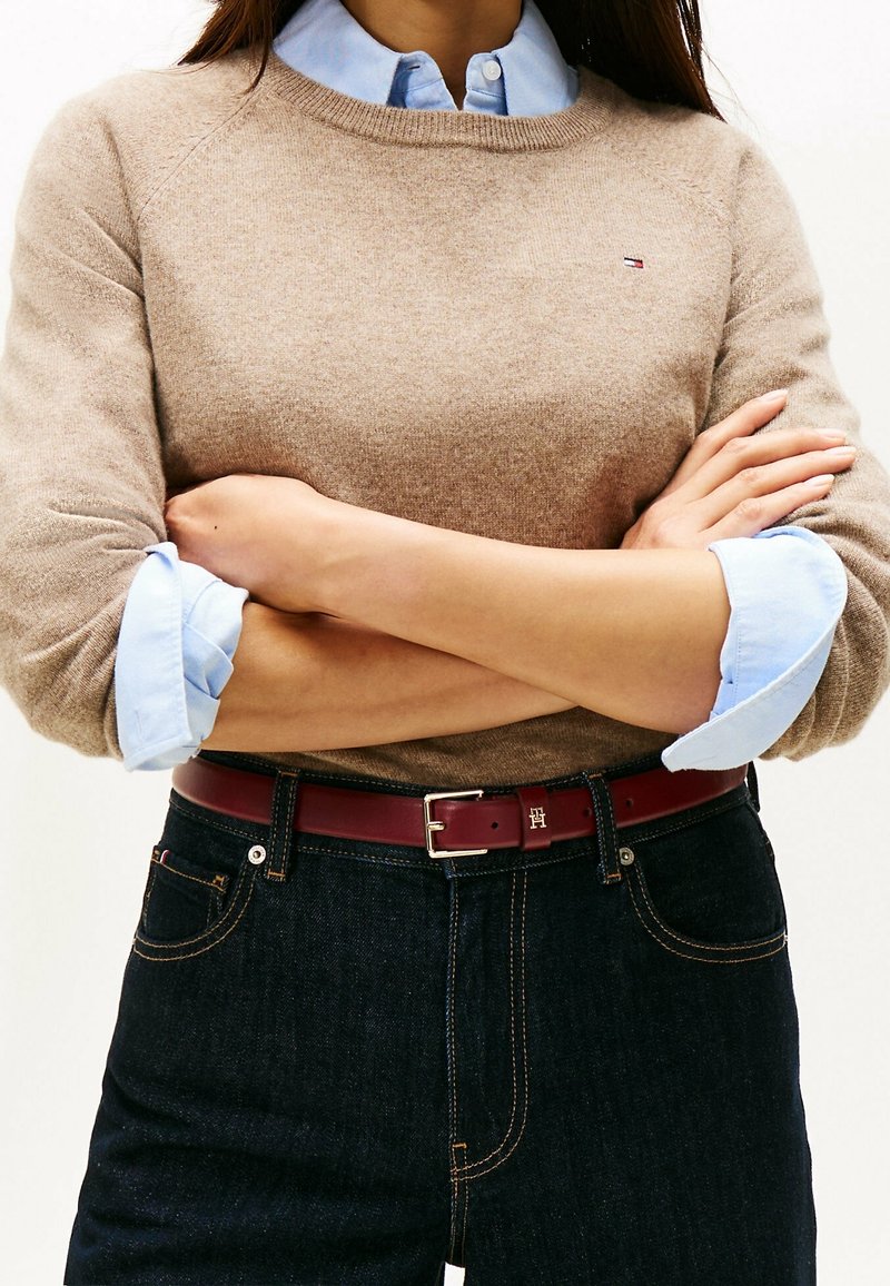 Femme les bras croisés portant un pull beige sur une chemise bleue à col, un jean foncé et une ceinture bordeaux avec une boucle métallique.
