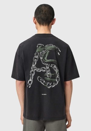 Homme portant un T-shirt noir avec un grand motif représentant un serpent vert enroulé de chaînes argentées dans le dos sur un fond uni.