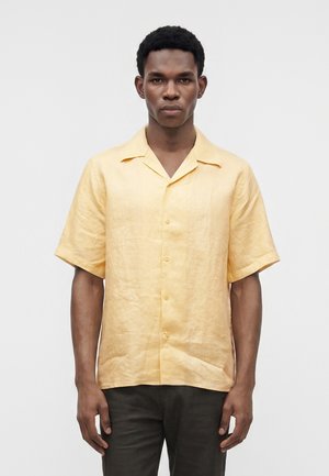 Homme portant une chemise jaune clair à manches courtes boutonnée, debout devant un fond gris clair uni.