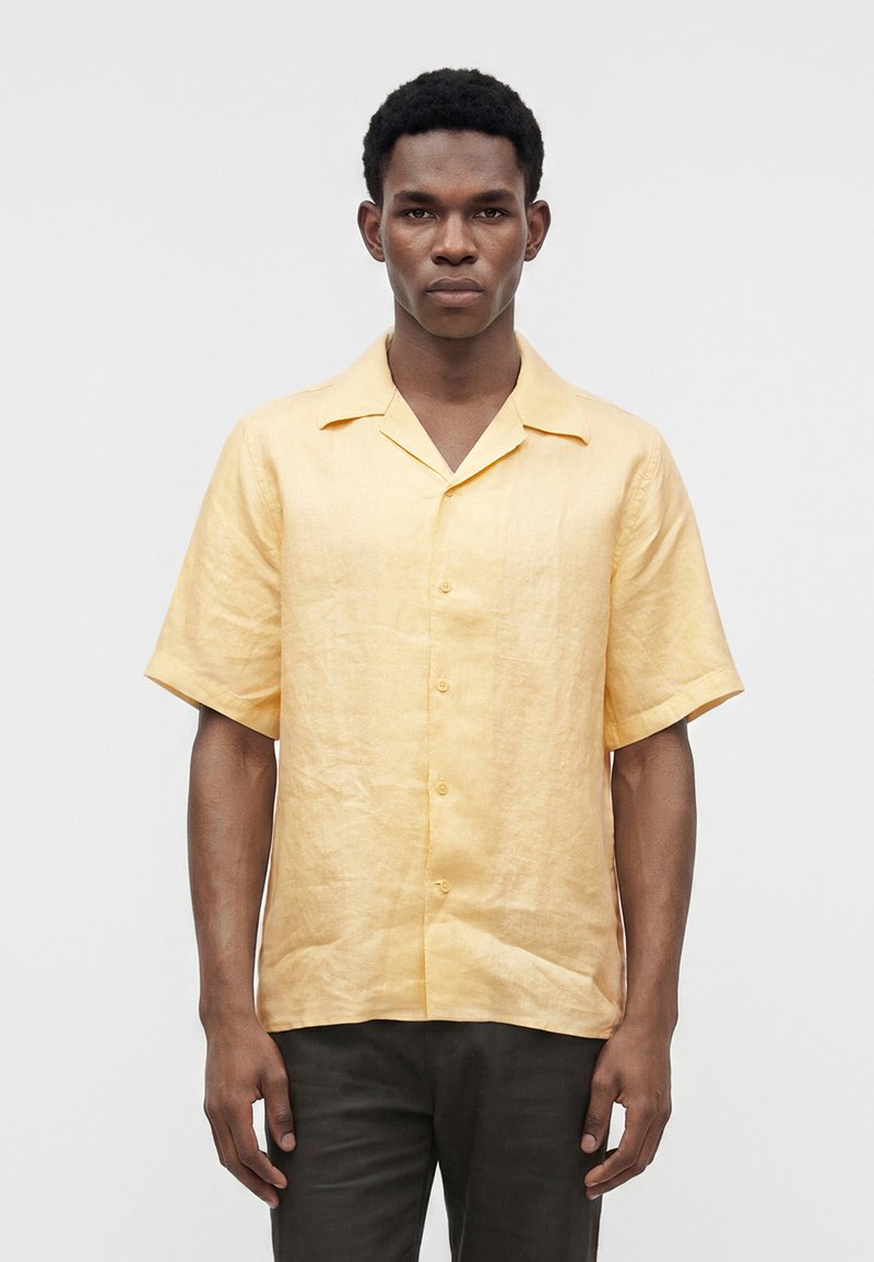 Homme portant une chemise jaune clair à manches courtes boutonnée, debout devant un fond gris clair uni.