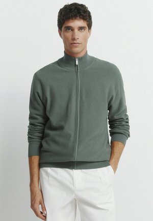 Felpa verde con zip e collo alto, polsini e orlo a coste. Il modello la indossa con pantaloni bianchi, evidenziando una vestibilità comoda e una texture distintiva.