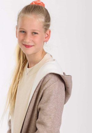 Hellbrauner Kapuzen-Cardigan über einem beigen Oberteil, mit einem weißen Innenfutter und einer glatten Textur. Lange blonde Haare, die mit einem orangefarbenen Haargummi zusammengebunden sind.
