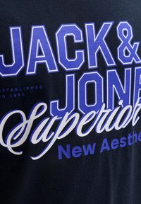 Črna bombažna majica s poudarjenim belim napisom "JACK & JONES" in "Superior" v vijolični barvi, spodaj pa "New Aesthetic". Standarden kroj z okroglim izrezom.