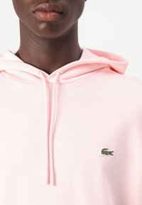 Sudadera de color rosa claro con cordones, que presenta un pequeño logo de cocodrilo verde de Lacoste en el lado izquierdo del pecho. Tejido suave y grosor medio.