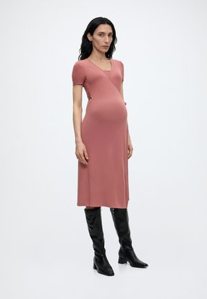 Tehotná žena v krátkych rukávoch, šaty po kolená v prašnatej ružovej farbe a čierne čižmy nad kolená na podpätku, stojaca pred jednoduchým bielym pozadím.