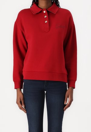 Czerwony, długi, z długim rękawem sweter z kołnierzykiem i trzema guzikami, noszony z ciemnoniebieskimi dżinsami, pokazany od szyi do bioder, ręce swobodnie opuszczone wzdłuż ciała.