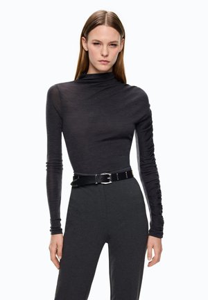 Donna che indossa un top nero aderente a maniche lunghe con arricciature e pantaloni grigio scuro a vita alta fissati da una cintura nera.