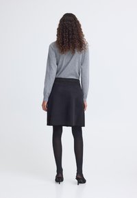 Frau mit lockigem Haar, die ein graues Langarmshirt, einen schwarzen Rock, schwarze Strumpfhosen und schwarze Absätze trägt, steht mit dem Rücken zur Kamera vor einem weißen Hintergrund.