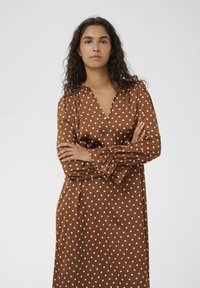 Rjava obleka z belo pikčasto vzorcem iz gladkega blaga, z V-izrezom in dolgimi, nabranimi rokavi ter sproščenim krojem.