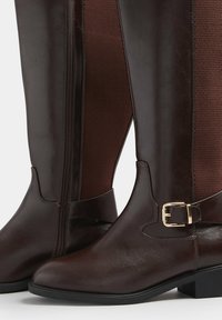 Bruna läderstövlar som når knäna, med en sidodragkedja, som har en strukturerad nätpanel och en guldfärgad spännrem som accent.