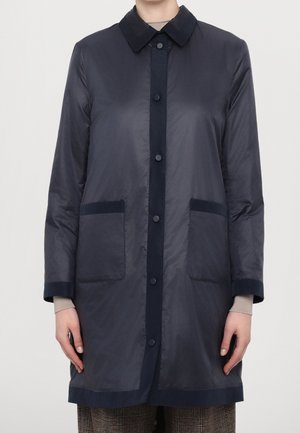 Femme portant un manteau long bleu marine à boutons avec deux poches avant, par-dessus un haut beige à manches longues et un pantalon à carreaux.
