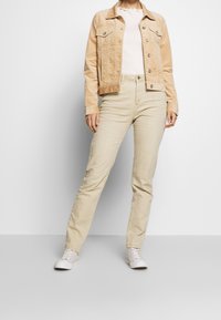 Chaqueta de pana beige con dos bolsillos en el pecho, combinada con pantalones ajustados beige claro y zapatillas blancas, presentando una tela texturizada.