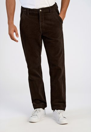 Mann trägt dunkelbraune Cordhosen und weiße Sneakers, mit einer Hand in der Tasche und weißem Hemd, das in die Hose gesteckt ist.