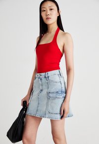 Rode getailleerde tanktop gecombineerd met een lichtblauwe denimrok met zijzakken, een rafelige zoom en een knoopsluiting. Zwarte tas in de hand.