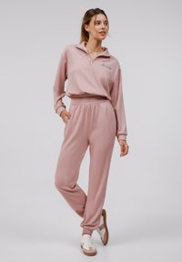 Pull-over zippé rose clair avec une petite poche avant et un pantalon de survêtement ajusté assorti. Tissu doux avec taille et poignets élastiques. Baskets blanches.