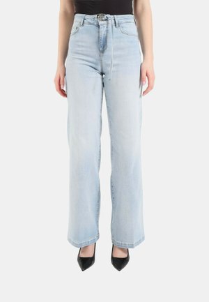 Donna che indossa jeans a gamba larga a vita alta azzurri chiari con cintura sottile e tacchi neri a punta, in piedi davanti a uno sfondo bianco.